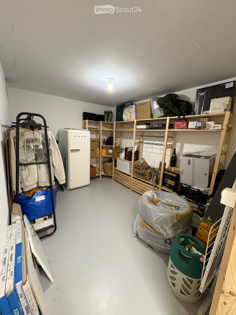A storage room with a refrigerator, shelves filled with boxes and items, a clothing rack, and various stored items including a backpack, laptop, and printer.