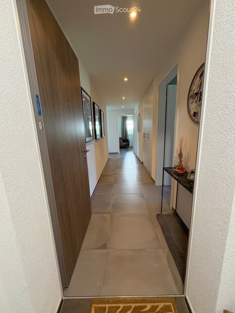 Clean, well-lit hallway with tiled flooring, white walls, and recessed lighting. Features a small console table with a vase and decorative items, framed artwork on the walls, and a glimpse of a living area at the end.