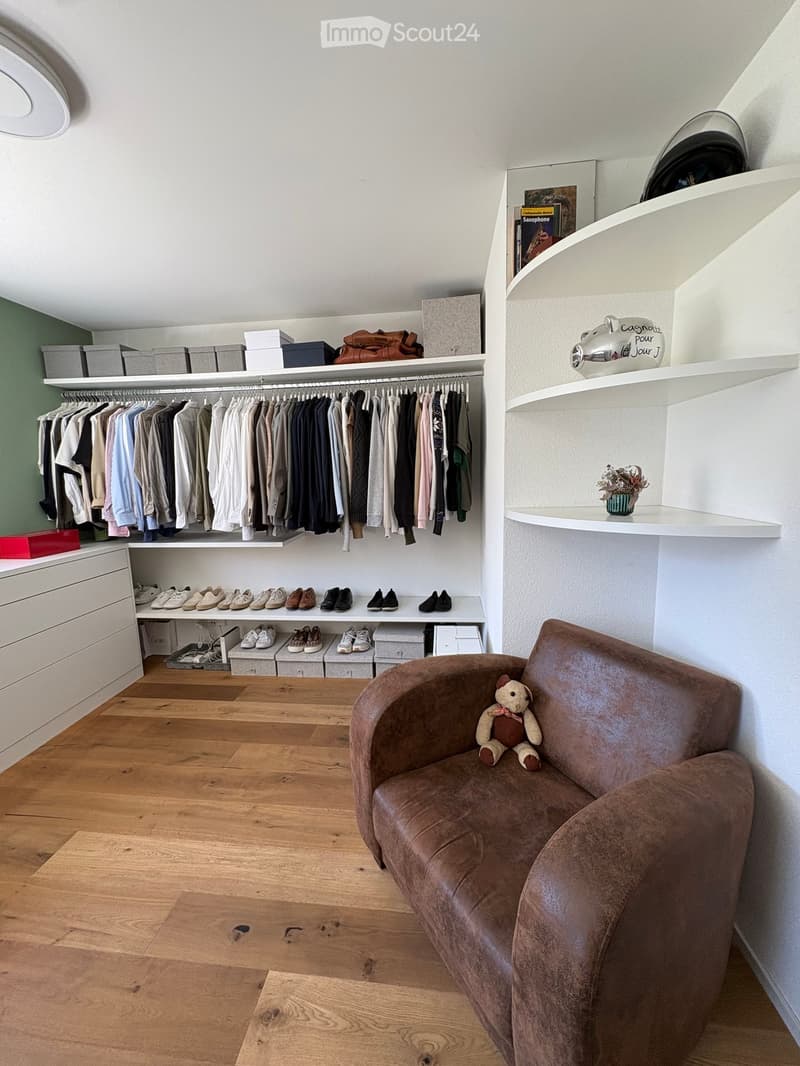 A well-organized walk-in closet with built-in shelves, a clothing rack, a shoe rack, and a small seating area featuring a brown leather sofa with a teddy bear. The room has wooden flooring and white walls.