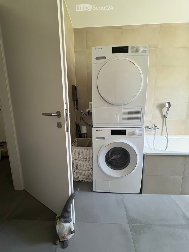 Bathroom with a stacked washing machine and dryer, a bathtub, a wicker basket, and a stuffed animal on the floor.