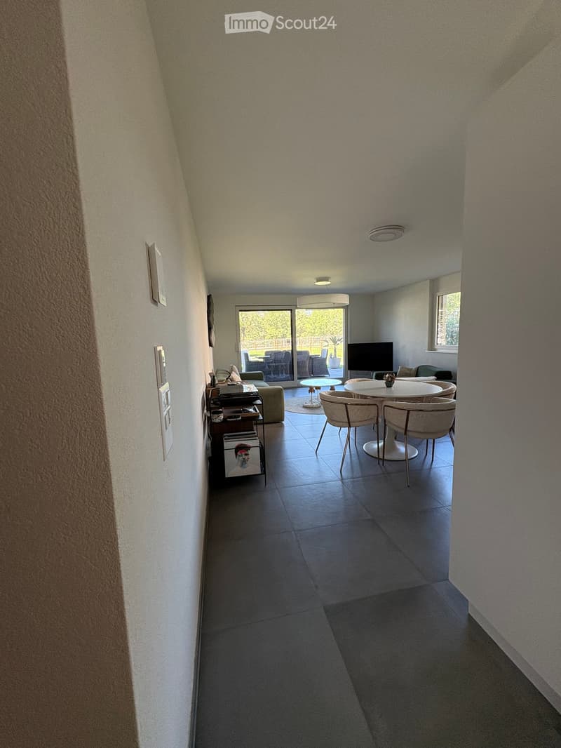 Open-plan living and dining area with a sofa, ottoman, dining table, chairs, television, and a small stool. The space features tiled flooring and large windows with a view of an outdoor patio.