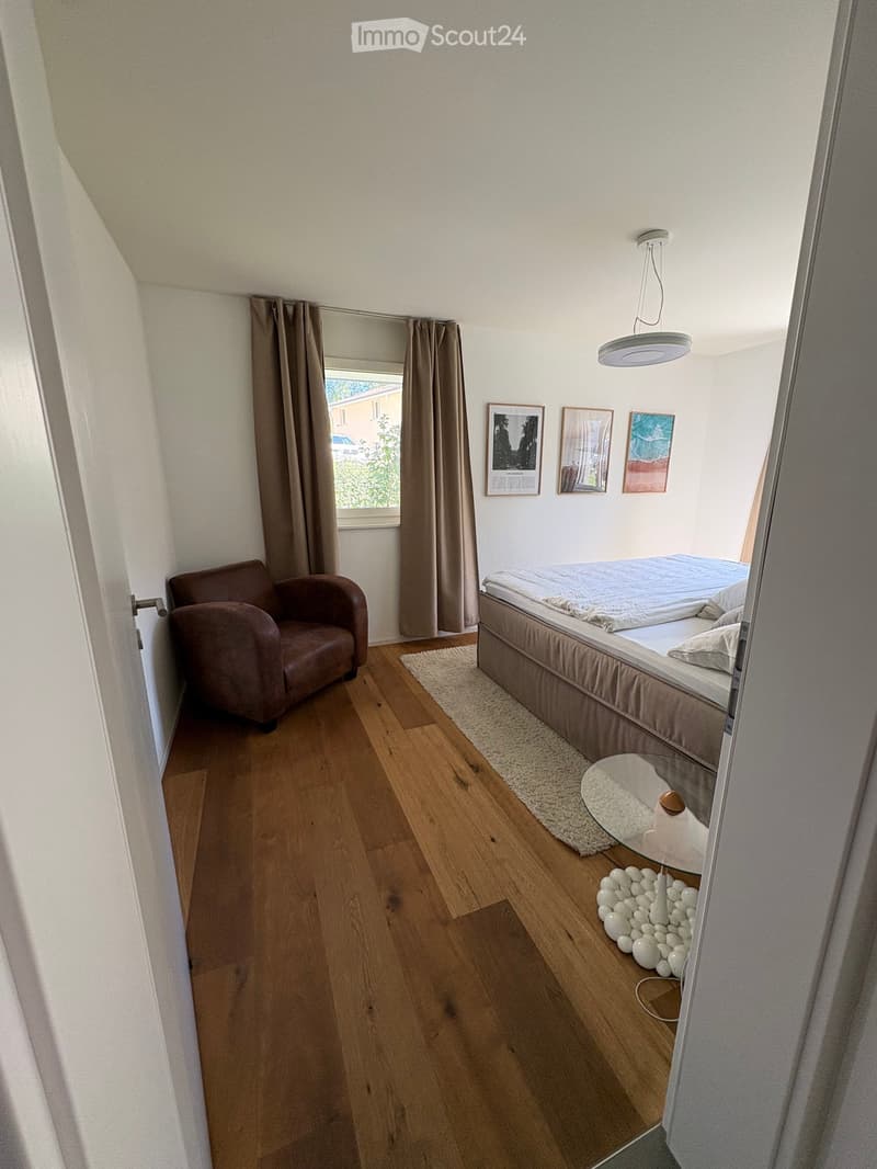 Cozy bedroom with wooden flooring, a bed with beige bedding, an armchair, a small table with a decorative arrangement, and framed pictures on the wall.