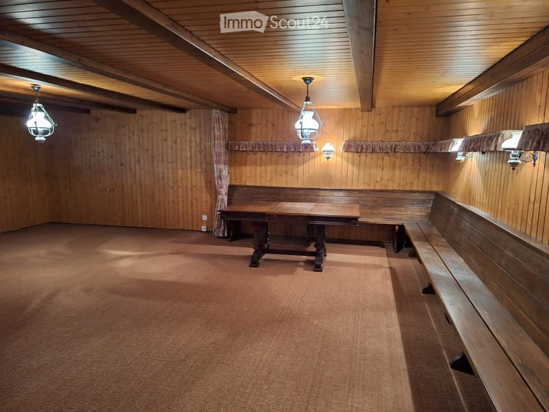 Wooden-paneled room with a central table and built-in benches, exposed wooden beams on the ceiling, and warm lighting from wall-mounted lamps.