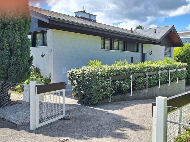 Two-story house with light blue exterior, grey roof, multiple windows, and a small chimney. The house is surrounded by a well-maintained hedge and a gravel driveway with a white gate.