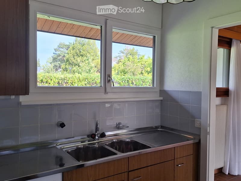 Kitchen with wooden cabinets, stainless steel sink, and tiled walls. Large windows provide natural light and a view of greenery outside.