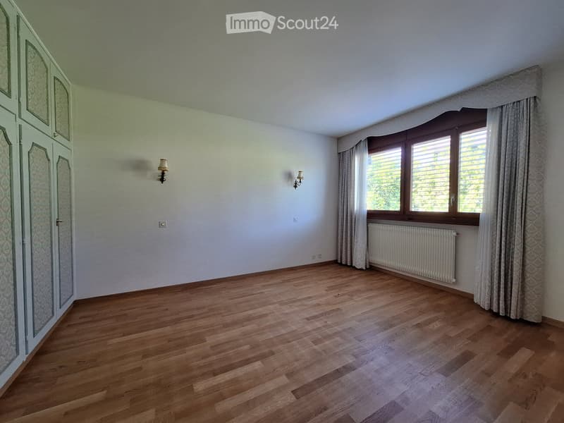 Empty room with wooden flooring, white walls, large window with curtains, radiator, and wall-mounted lamps.