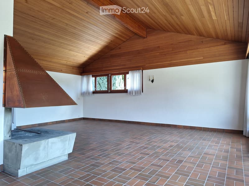 Empty room with wooden ceiling, brick floor, white walls, and a fireplace with a copper hood. Windows with white curtains are visible.