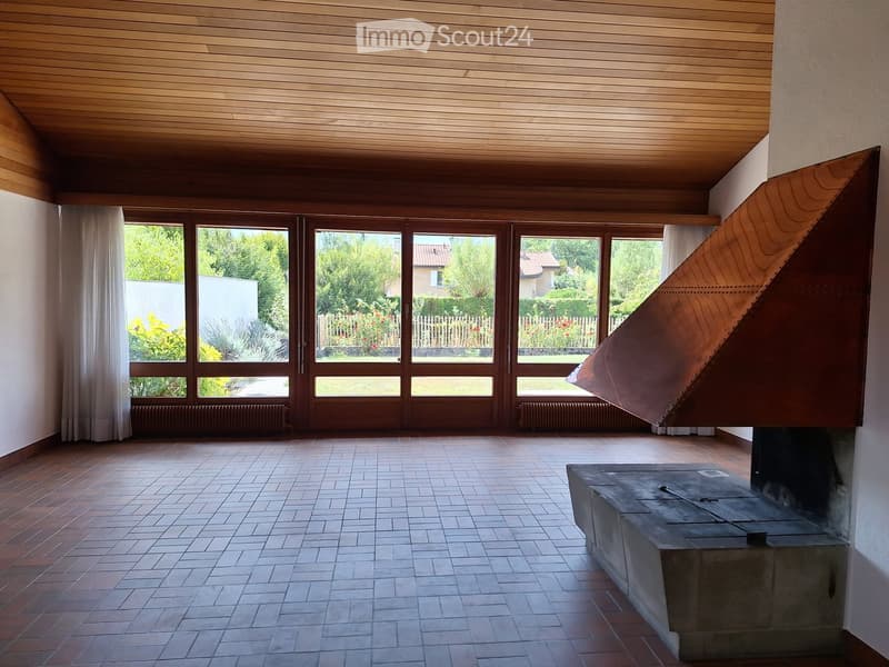 An empty room with wooden ceiling, large windows, and a fireplace. The space features a tiled floor and wooden walls.