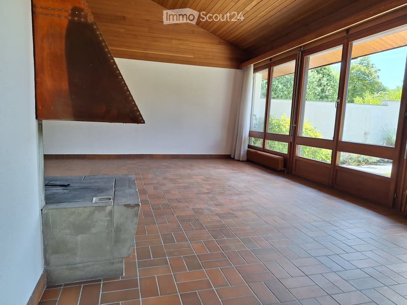 An unfurnished room with a tiled floor, large windows, and a concrete fireplace. The room features wooden ceiling beams and a radiator under the windows.