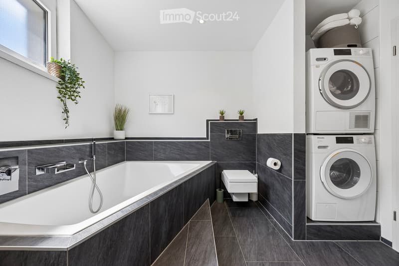 Modern bathroom with a bathtub, toilet, washing machine, dryer, and potted plants. Features dark grey tiles and a sleek design.