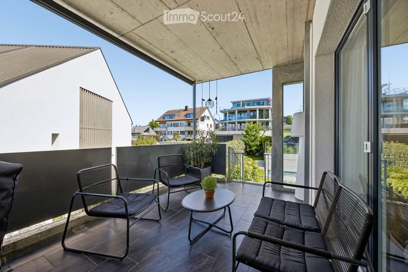 Modern balcony with dark wooden floor, black metal chairs, a bench, a small round table, and potted plants. The balcony has a wooden ceiling and glass walls, offering a view of the surrounding area.