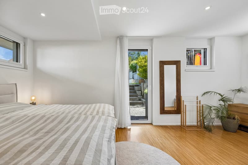 Bright bedroom with wooden flooring, large bed with striped bedding, a wooden-framed mirror, a small table lamp, and a potted plant near the window. A sliding glass door leads to an outdoor staircase.