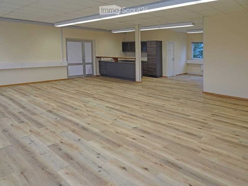 Empty room with wooden flooring, white walls, and a small kitchen area in the background.
