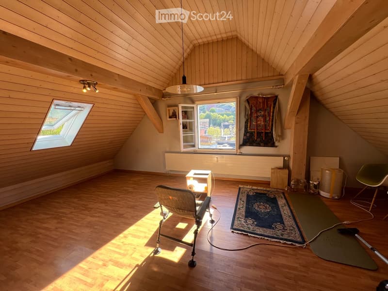 Bright attic room with wooden walls and ceiling, large windows, a chair, small table, rug, yoga mat, and storage items.