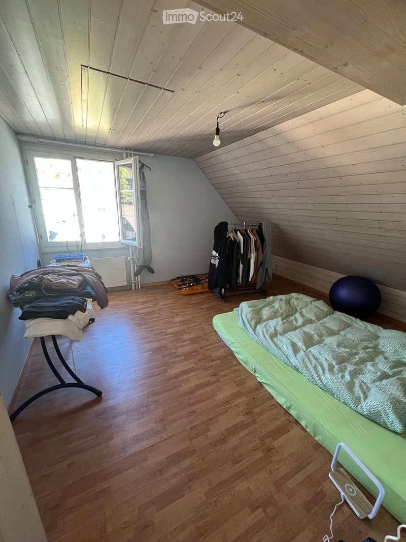 Bedroom with wooden floor, slanted wooden ceiling, two beds, clothing rack, and a large window with open shutters.