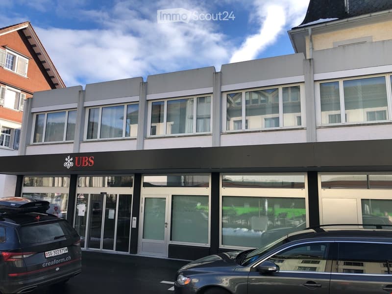 Modern commercial building with large glass windows and a black sign reading "UBS" above the entrance. Two cars are parked in front.