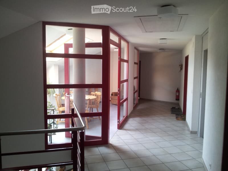 A well-lit hallway with tiled flooring, white walls, and red-framed glass doors leading to a room with wooden chairs and a table. A fire extinguisher is mounted on the wall.