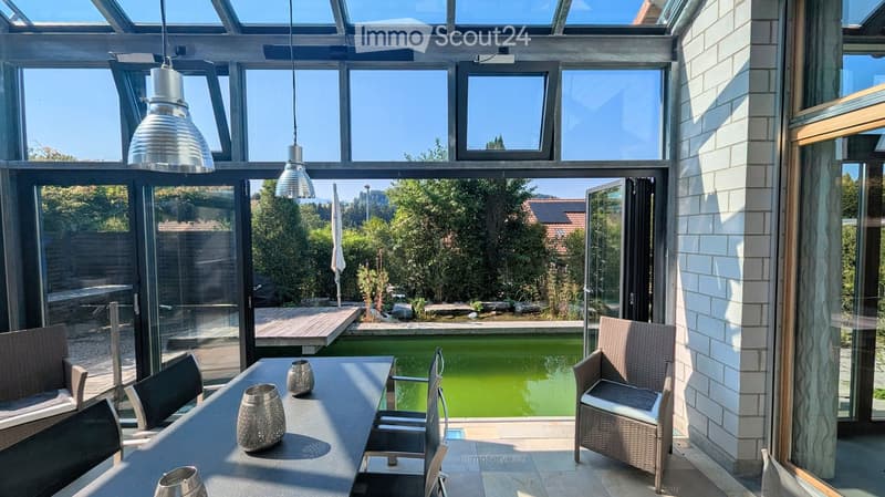 Bright, modern room with large glass windows and a view of a swimming pool and garden. Features a table with chairs and decorative lamps.