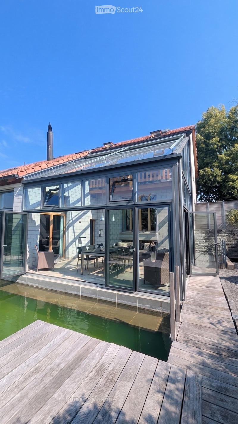 Modern outdoor area with a swimming pool, wooden decking, and a glass-enclosed patio connected to the house. The patio features a dining table and chairs, and the house has a red-tiled roof and white brick walls.