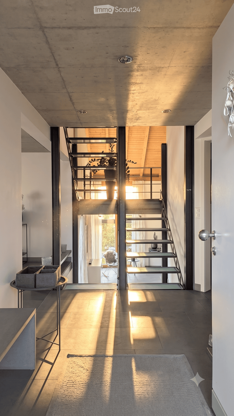 Modern hallway with a staircase leading upwards, featuring a sleek metal railing, wooden steps, and natural light streaming in from the adjacent living area.