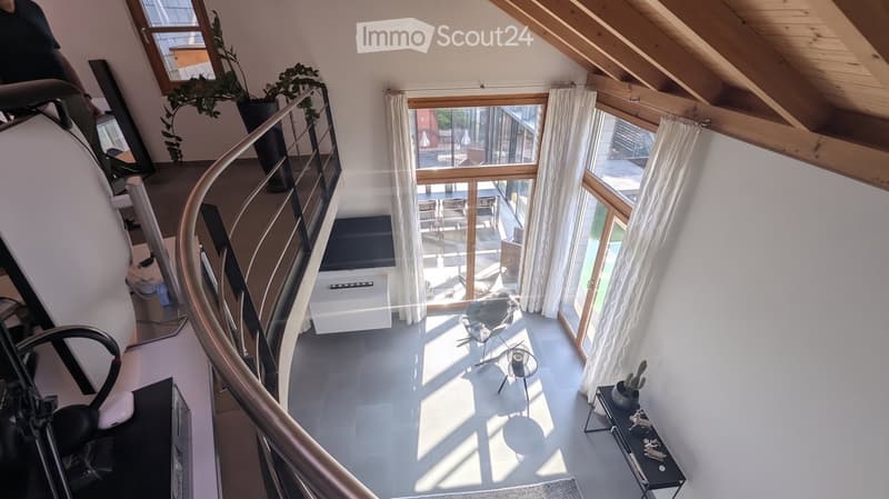 Modern living room with white walls, wooden ceiling beams, large windows with white curtains, and a spiral staircase leading to an upper level. The room features a sleek black chair, a small round table, and a TV screen mounted on the wall.