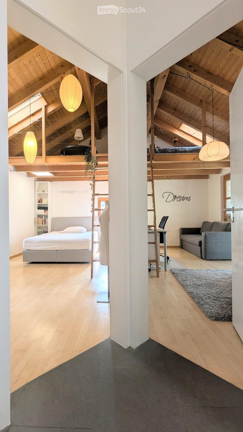 Open-plan living and bedroom area with wooden floors, exposed wooden beams, pendant lights, a sofa, a chair, a table with a laptop, a bed, and a bookshelf.