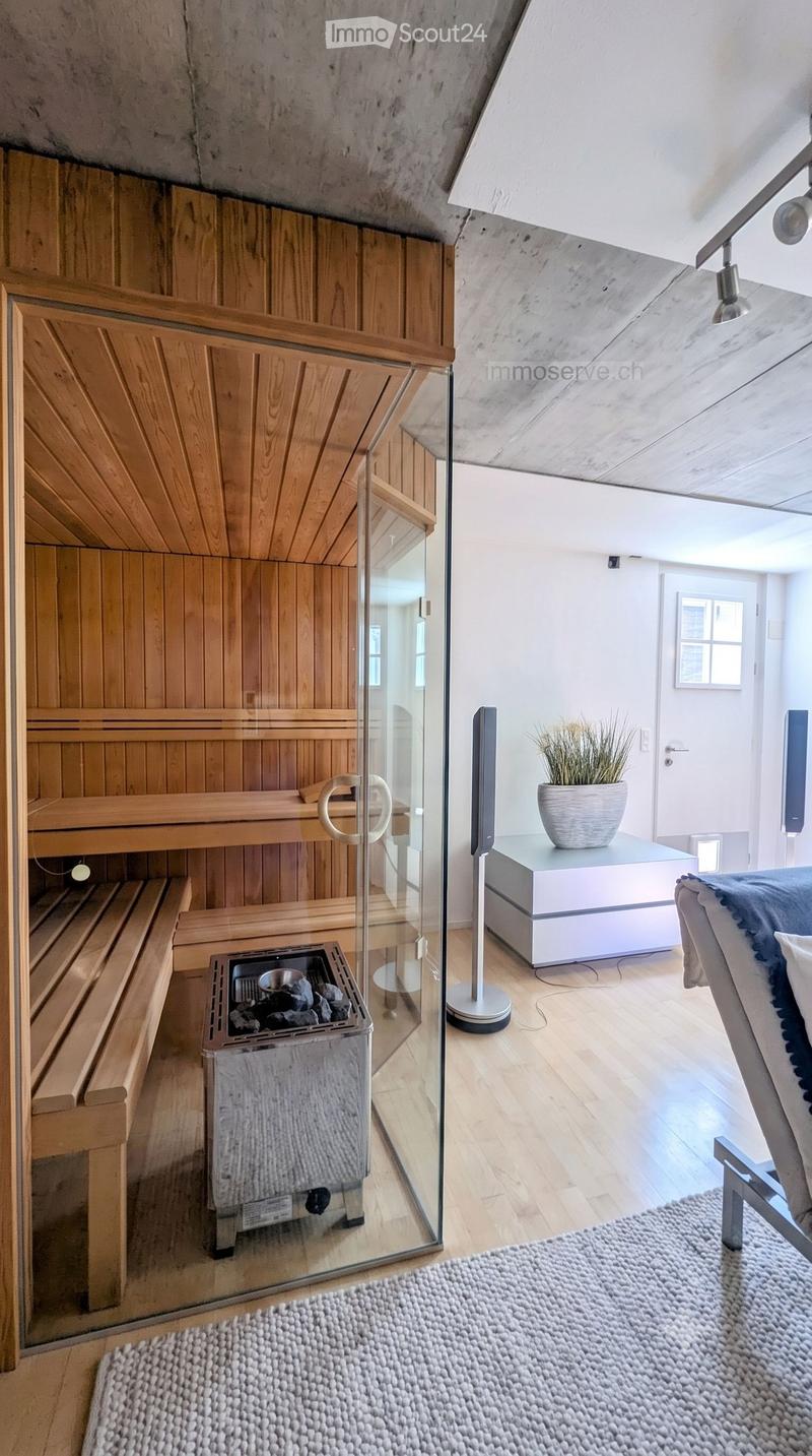 A modern sauna room with wooden benches, a glass door leading to a living space, a small table with a potted plant, a chair, and a rug on the floor.