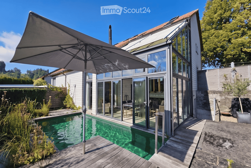 A modern outdoor area features a swimming pool, large glass doors leading to an indoor space, a patio umbrella, and surrounding greenery.