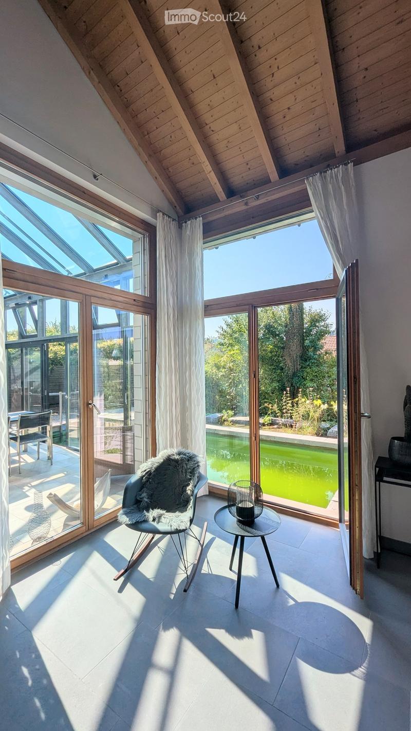 Bright, modern room with large windows and a skylight, featuring a fur-covered chair, small round table, and decorative vase. The room has a swimming pool visible through the windows.