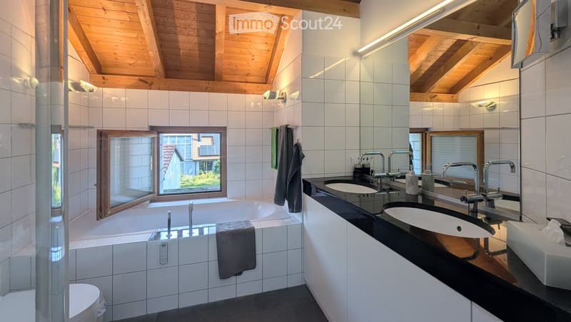Modern bathroom with a large bathtub, double sink vanity, and wooden ceiling beams. Features include a large window, towels, and a toilet.