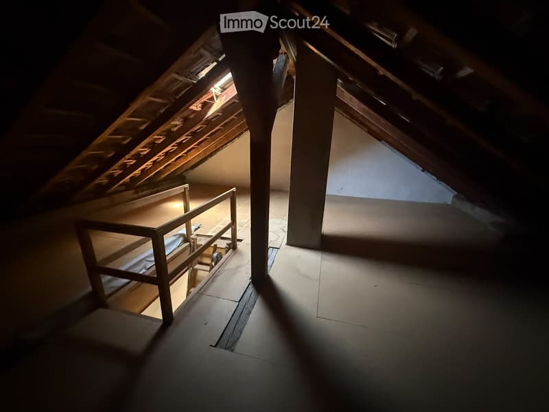 Empty attic space with wooden beams, exposed rafters, and a small window letting in natural light. The floor is concrete, and there is a wooden railing on one side.