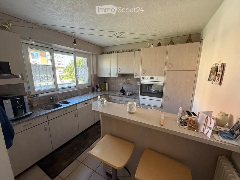 Modern kitchen with light wood cabinets, granite countertops, and stainless steel appliances. Features a central island with bar stools, a sink, and a coffee maker on the counter.