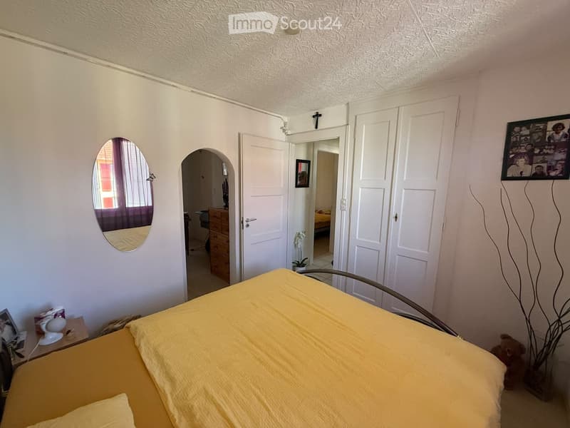 Bedroom with a yellow bedspread, a nightstand with a lamp, a large mirror, a wardrobe, and a picture frame on the wall. An open door leads to another room.