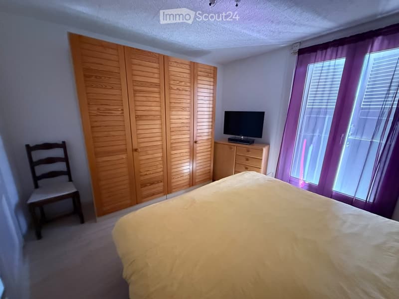 Bedroom with a large bed, wooden wardrobe, chair, television on a small cabinet, and large window with purple curtains.