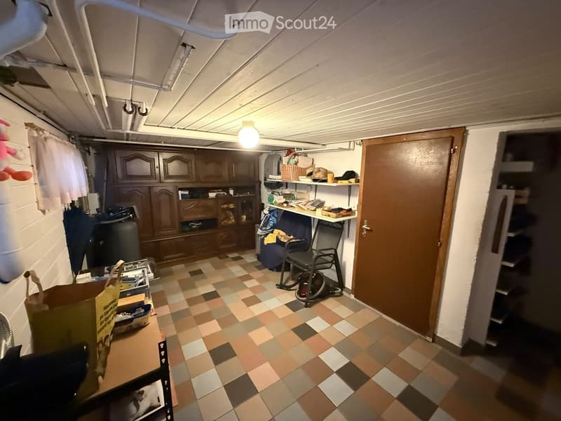 A cluttered basement or storage room with a tiled floor, wooden cabinet, shelves, a chair, and various items scattered around.