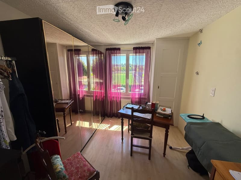 A well-lit room with wooden flooring, a large mirror, a wardrobe, a small table with a chair, and an ironing board with an iron on it.