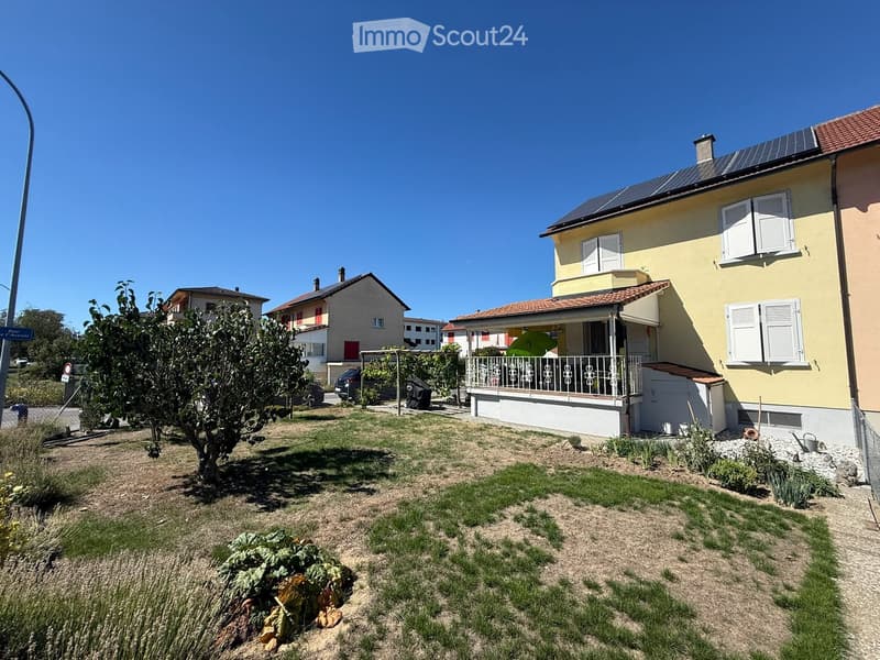 A two-story yellow house with white shutters and a balcony, surrounded by a small garden with grass, plants, and a tree.