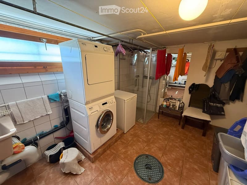 Bathroom with a washing machine and dryer, a small shower area, a stool, a chair, a small table, and various towels and items.