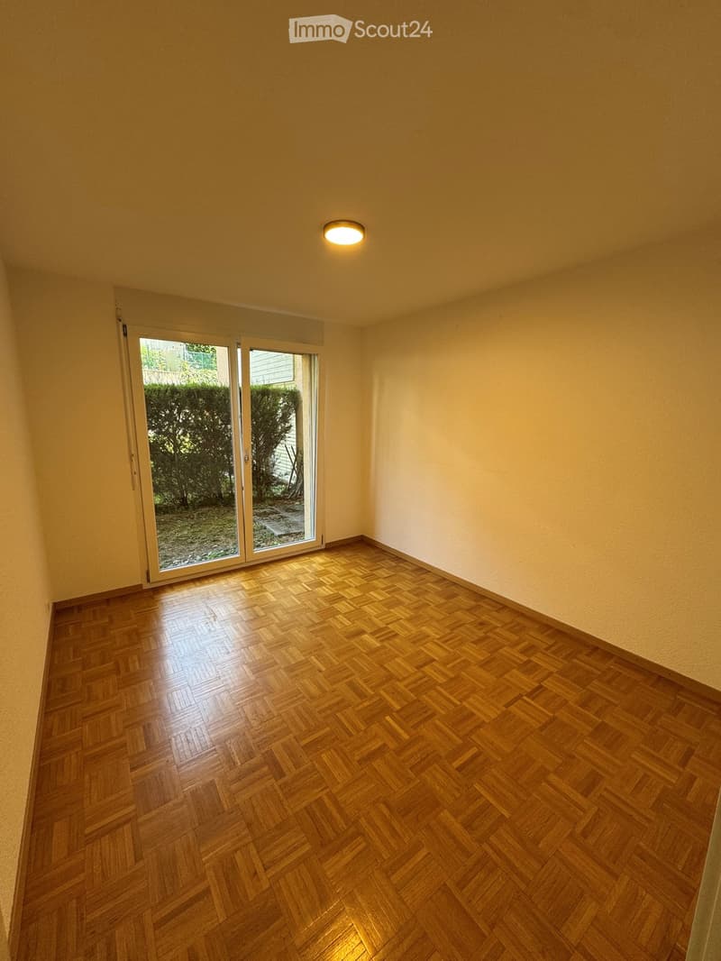 Empty room with wooden herringbone flooring, white walls, and a large sliding glass door leading to a garden area.