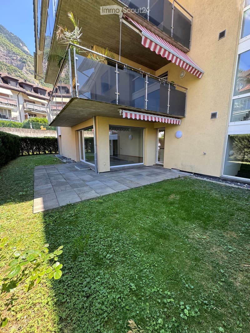 A well-maintained garden area with a tiled patio, green grass, and a modern house with balconies and striped awnings.