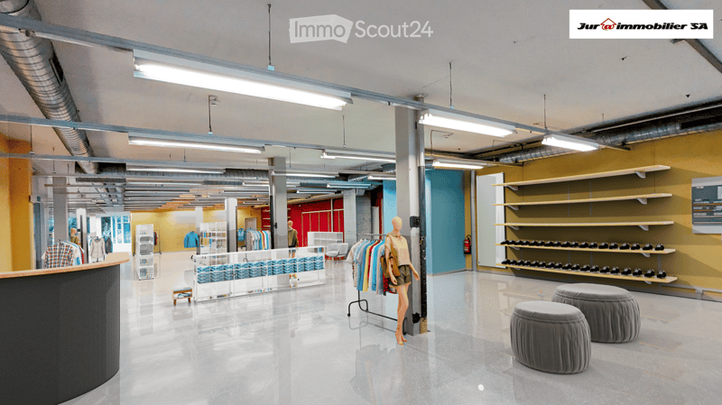 Modern retail space with polished concrete floors, exposed ceiling beams, and bright lighting. Features include grey stools, clothing racks, shelves, and a mannequin.
