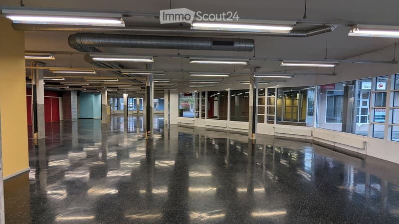 Empty, wide corridor with polished black flooring, large windows on one side, and exposed ceiling pipes and lighting fixtures.