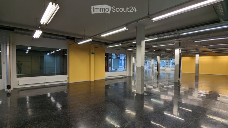 Empty, spacious room with polished black flooring, large windows, and exposed structural columns. The walls are painted in a bright yellow color, and the ceiling features industrial-style lighting fixtures.