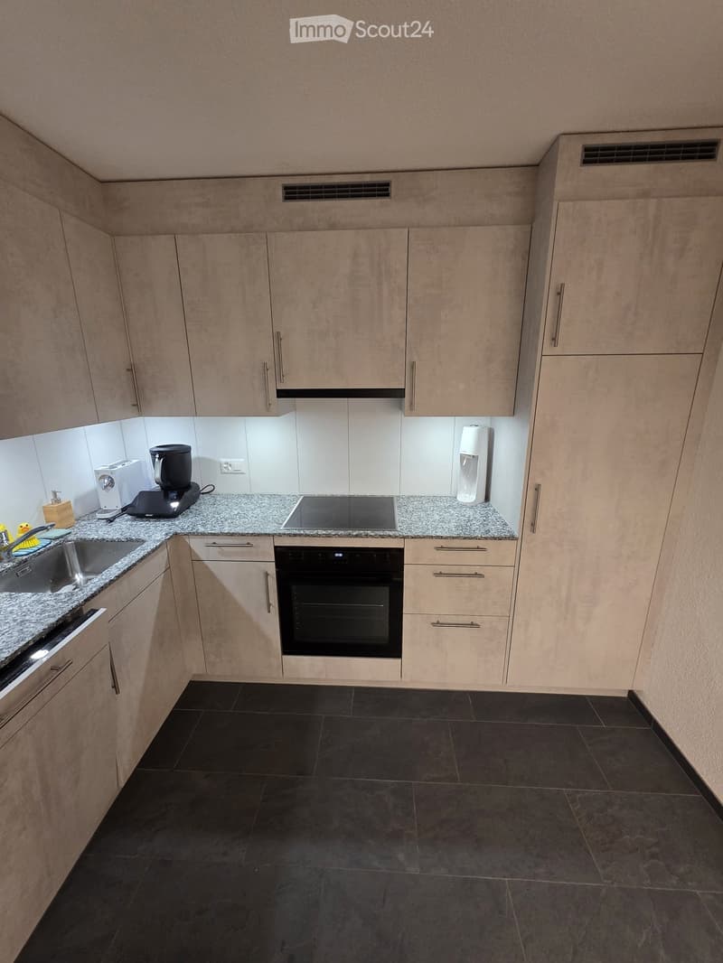 Modern kitchen with light wood cabinets, granite countertops, and black tiled floor. Features include a sink, oven, coffee maker, and water bottle on the counter.