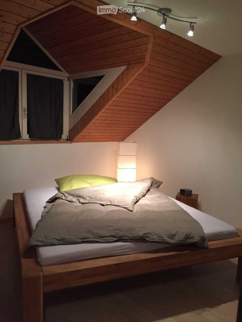 Cozy bedroom with wooden slanted ceiling, large window with black curtains, neatly made bed with pillows and duvet, nightstand with lamp, and modern track lighting.