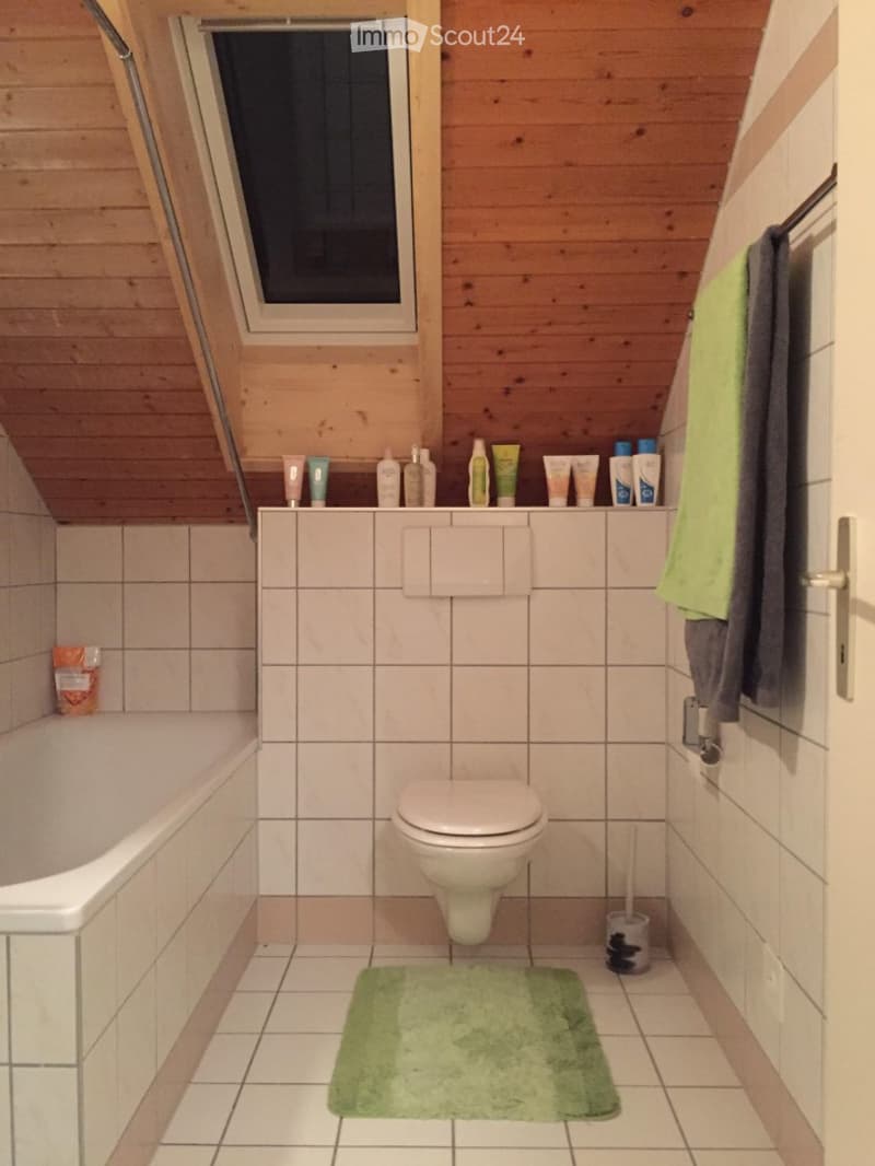 Cozy bathroom with wooden slanted ceiling, white tiled walls and floor, bathtub, toilet, towel, and cosmetics on the shelf.