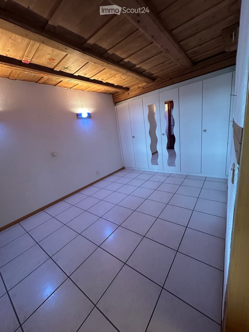 Empty room with tiled floor, white walls, and built-in white cabinets with mirrored doors. Exposed wooden ceiling beams and a small wall-mounted light fixture are visible.