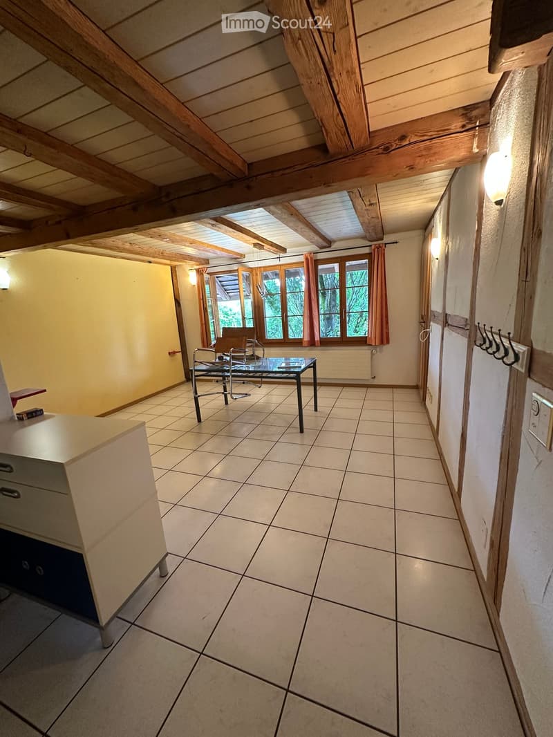 Bright, open room with exposed wooden beams, tiled floor, large windows with curtains, and a small table with chairs.
