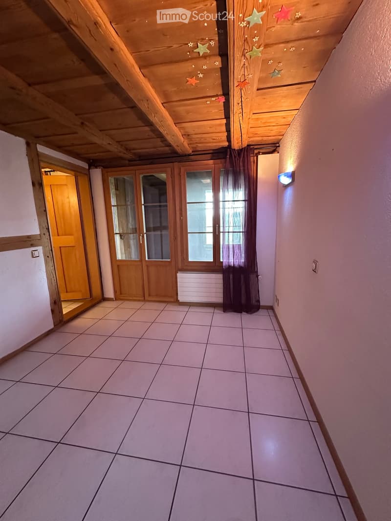 Empty hallway with tiled floor, wooden doors, and exposed wooden beams on the ceiling. Features a wall-mounted light fixture and a radiator.