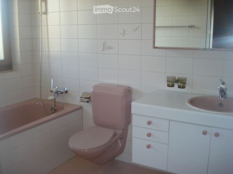 A small bathroom with white tiled walls and a pink bathtub, toilet, and sink. A white cabinet is positioned next to the sink, and a mirror is mounted on the wall above it.